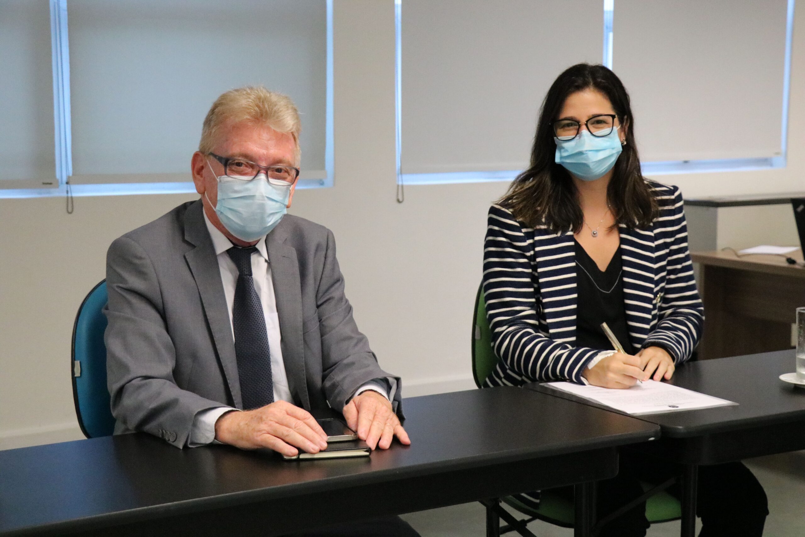Na foto estão sentados lado a lado e de máscara, posando para a câmera, o presidente do CRMV-SP e a médica-veterinária Brana Sanctos, que faz a menção de assinar o termo de posse como integrante da Comissão de Nutrição Animal.