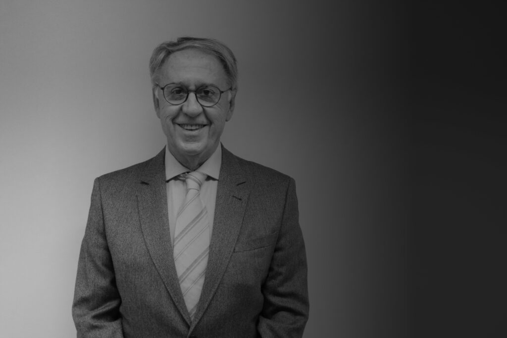 A imagem está em preto e branco e traz a foto do professor Carlos Eduardo Larsson, um senhor de óculos, sorridente, vestindo terno e gravata.