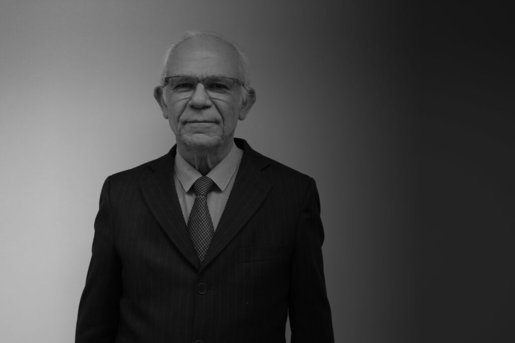 A imagem em preto e branco traz a foto do professor Silvio Arruda Vasconcellos, um senhor de óculos, vestindo terno e gravata.