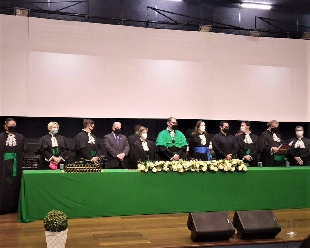 Na foto aparece a mesa de honra composta da solenidade de colação de grau, da qual o vice-presidente do CRMV-SP faz parte, ao lado do diretor, da vice-diretora e de docentes da FMVZ-USP. A mesa está decorada com uma toalha verde, flores brancas e os canudos dos diplomas que serão entregues.