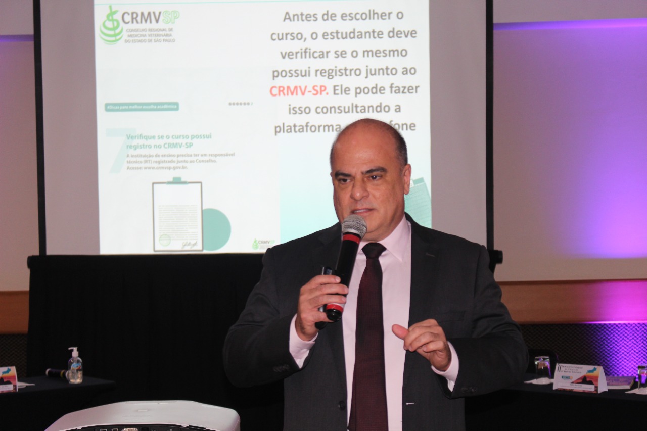 Presidente da Comissão Técnica de Educação do CRMV-SP apresenta ao microfone o projeto Melhor Escolha do CRMV-SP. No telão atrás dele está o slide tratando das ações de São Paulo na área da Educação.