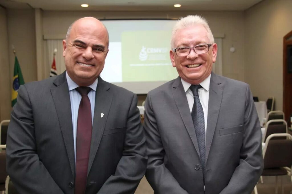 Presidente e vice-presidente do CRMV-SP posam juntos vestindo terno e gravata. Ambos estão sorrindo e dispostos em frente a um telão com a logomarca do CRMV-SP sob um fundo verde.