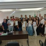 Novos profissionais que receberam a cédula de identidade posam juntos em pé mostrando os novos documentos.