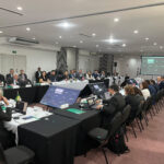 Foto mostra os participantes sentados, dispostos com mesas à frente de cada um, em formato quadrado.