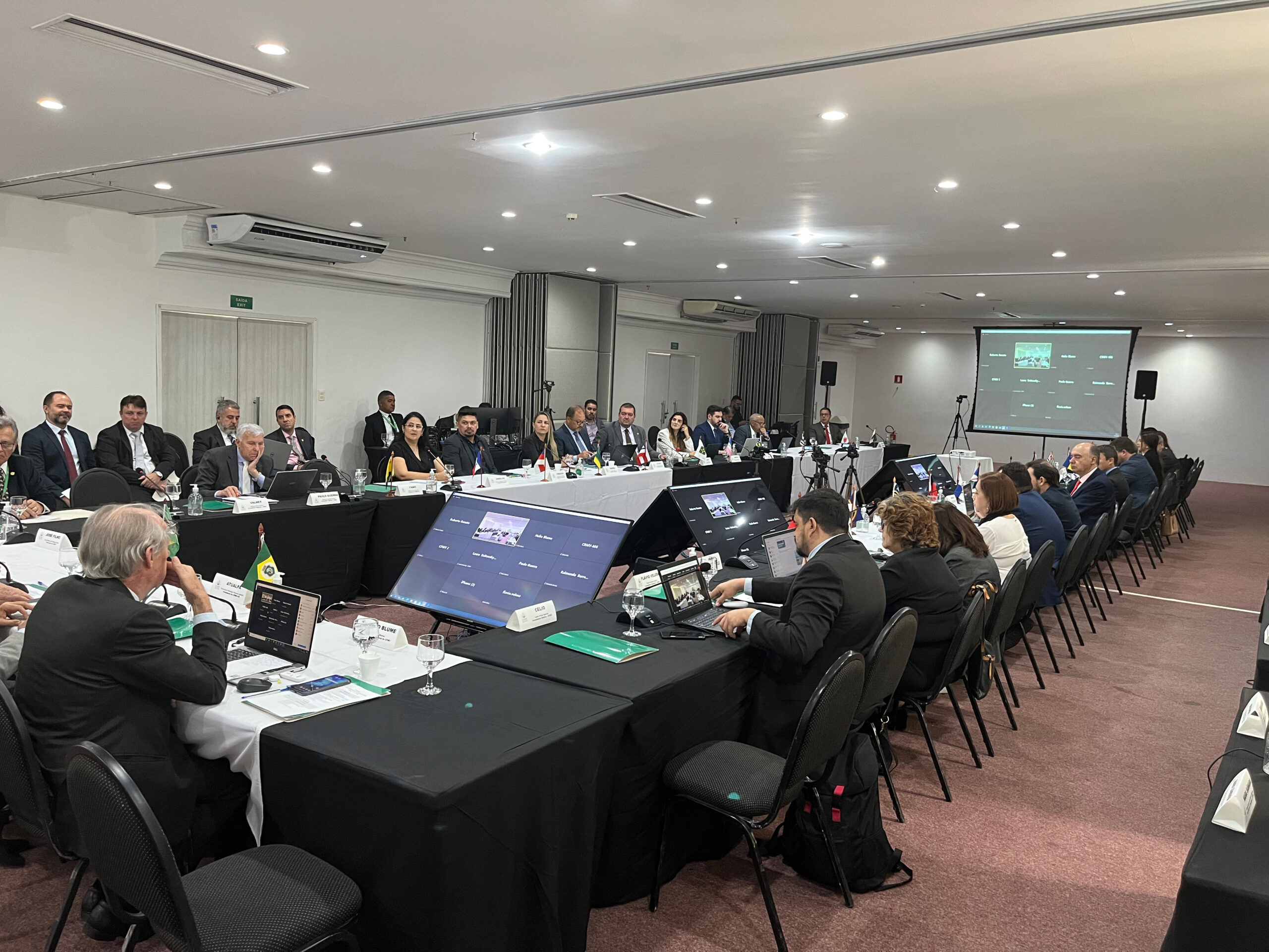 Foto mostra os participantes sentados, dispostos com mesas à frente de cada um, em formato quadrado.