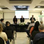 A foto foi tirada da parte de trás do auditório e mostra os participantes sentados em cadeiras de costas para a câmera. A frente estão posicionados atrás de uma mesa com toalha preta e um arranjo de flores, a tesoureira, o presidente e o secretário-geral do CRMV-SP. Eles estão de pé e ao microfone está o secretário-geral.