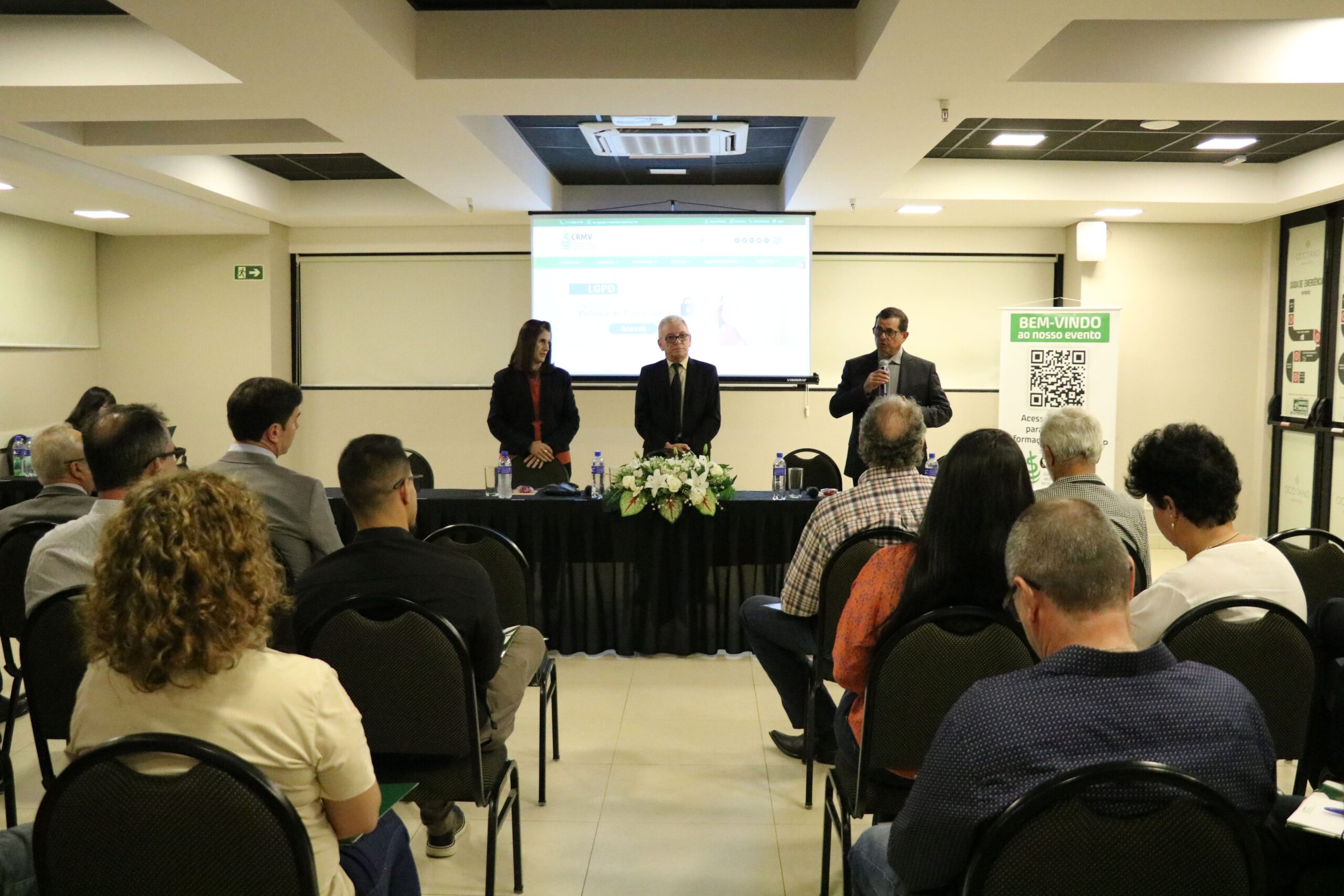 A foto foi tirada da parte de trás do auditório e mostra os participantes sentados em cadeiras de costas para a câmera. A frente estão posicionados atrás de uma mesa com toalha preta e um arranjo de flores, a tesoureira, o presidente e o secretário-geral do CRMV-SP. Eles estão de pé e ao microfone está o secretário-geral.
