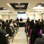 A foto foi tirada da parte de trás do auditório e mostra os participantes sentados em cadeiras de costas para a câmera. A frente estão posicionados atrás de uma mesa com toalha preta e um arranjo de flores, a tesoureira, o presidente e o secretário-geral do CRMV-SP. Eles estão de pé e ao microfone está o secretário-geral.