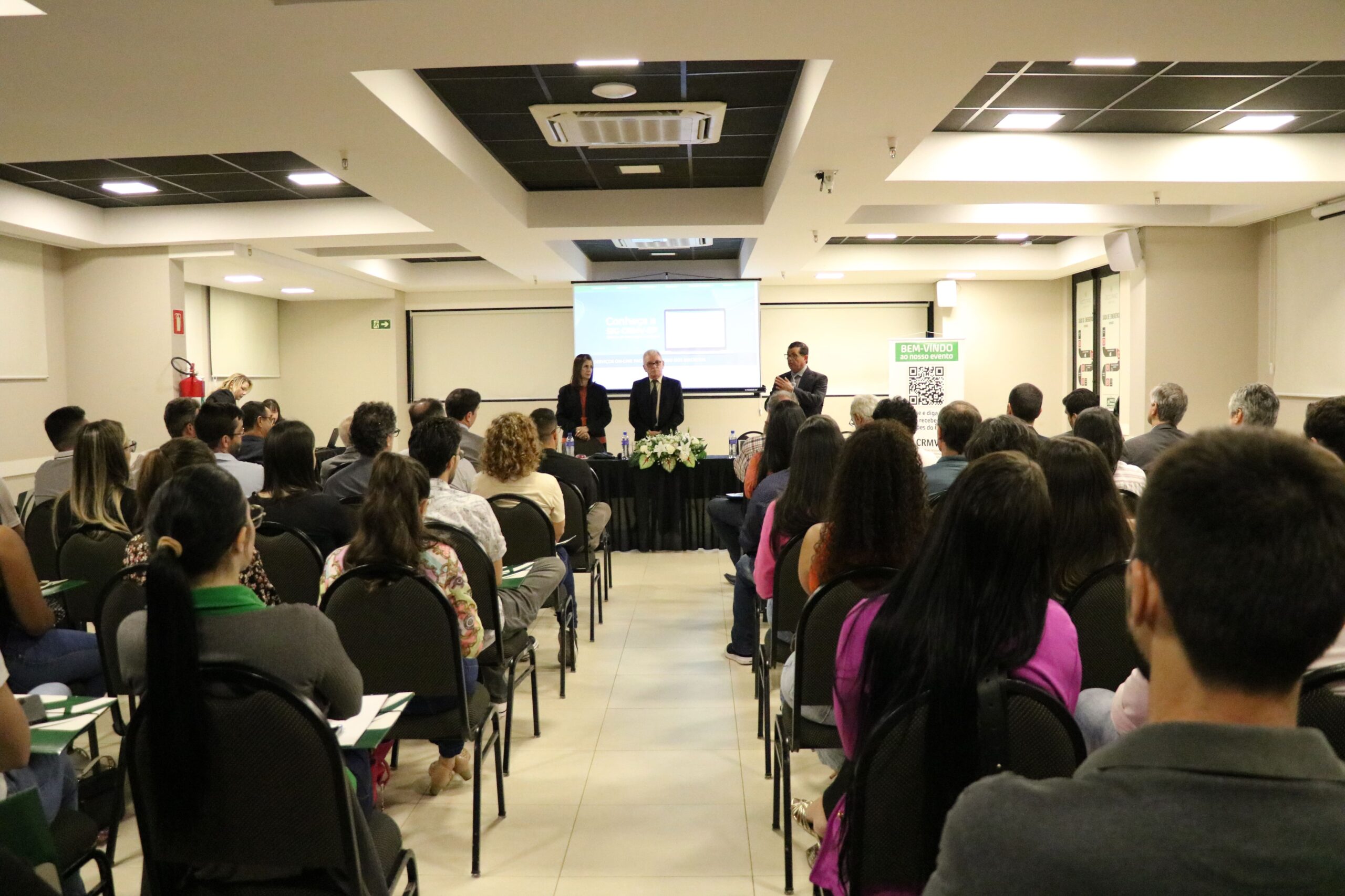 A foto foi tirada da parte de trás do auditório e mostra os participantes sentados em cadeiras de costas para a câmera. A frente estão posicionados atrás de uma mesa com toalha preta e um arranjo de flores, a tesoureira, o presidente e o secretário-geral do CRMV-SP. Eles estão de pé e ao microfone está o secretário-geral.