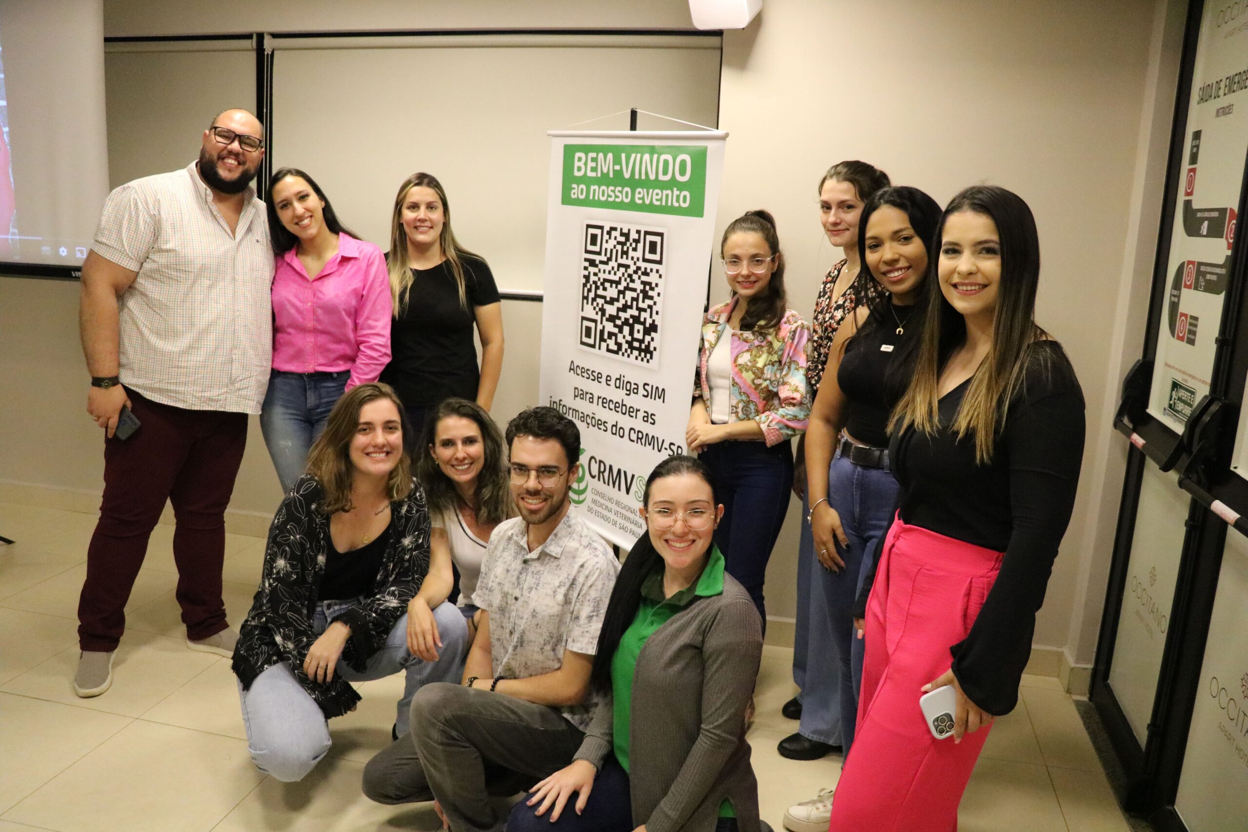 Na foto aparecem o coordenador de curso da Unimep e alunos da graduação. Eles estão posando e sorriem para a câmera. Estão posicionados parte de pé e parte agachados. Ao centro está o banner do CRMV-SP com a frase "Bem-vindos ao nosso evento".