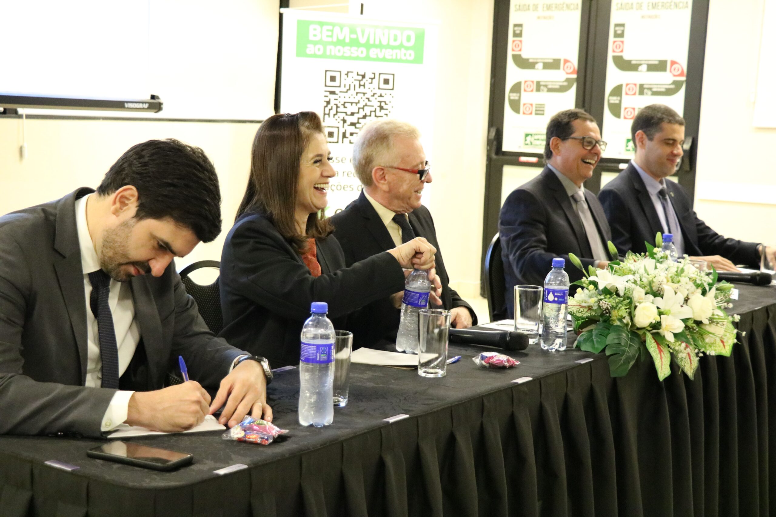 A foto mostra mesa com toalha preta e arranjo de flores composta pelo assessor jurídico, pela tesoureira, pelo presidente, pelo secretário-geral e pelo diretor técnico do CRMV-SP. Eles sorriem de maneira descontraída. O assessor jurídico faz anotações.