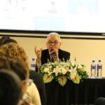 A foto foi tirada da parte de trás do auditório e mostra os participantes sentados em cadeiras de costas para a câmera. A frente estão posicionados atrás de uma mesa com toalha preta e um arranjo de flores, o presidente e o secretário-geral do CRMV-SP. Eles estão sentados. A imagem dá foco maior ao presidente do CRMV-SP falando ao microfone.