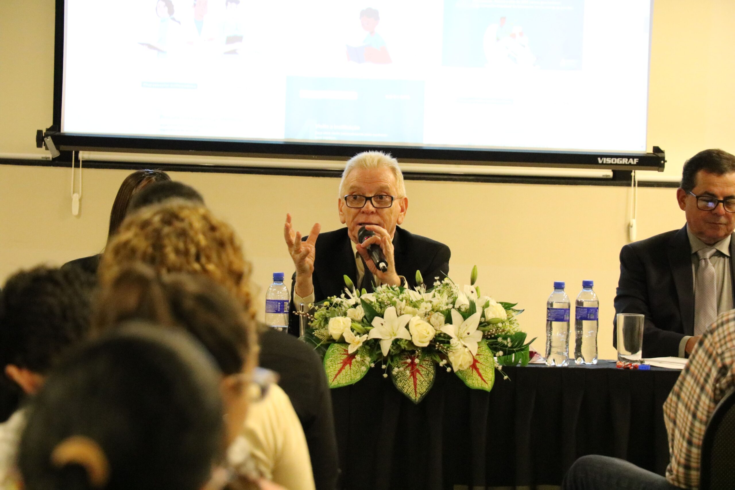 A foto foi tirada da parte de trás do auditório e mostra os participantes sentados em cadeiras de costas para a câmera. A frente estão posicionados atrás de uma mesa com toalha preta e um arranjo de flores, o presidente e o secretário-geral do CRMV-SP. Eles estão sentados. A imagem dá foco maior ao presidente do CRMV-SP falando ao microfone.
