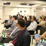 A foto mostra o auditório do CRMV-SP Escuta de Piracicaba lotado de pessoas sentadas em cadeiras com pastas do CRMV-SP verde e brancas no colo. A frente está de pé ao microfone o presidente do CRMV-SP.