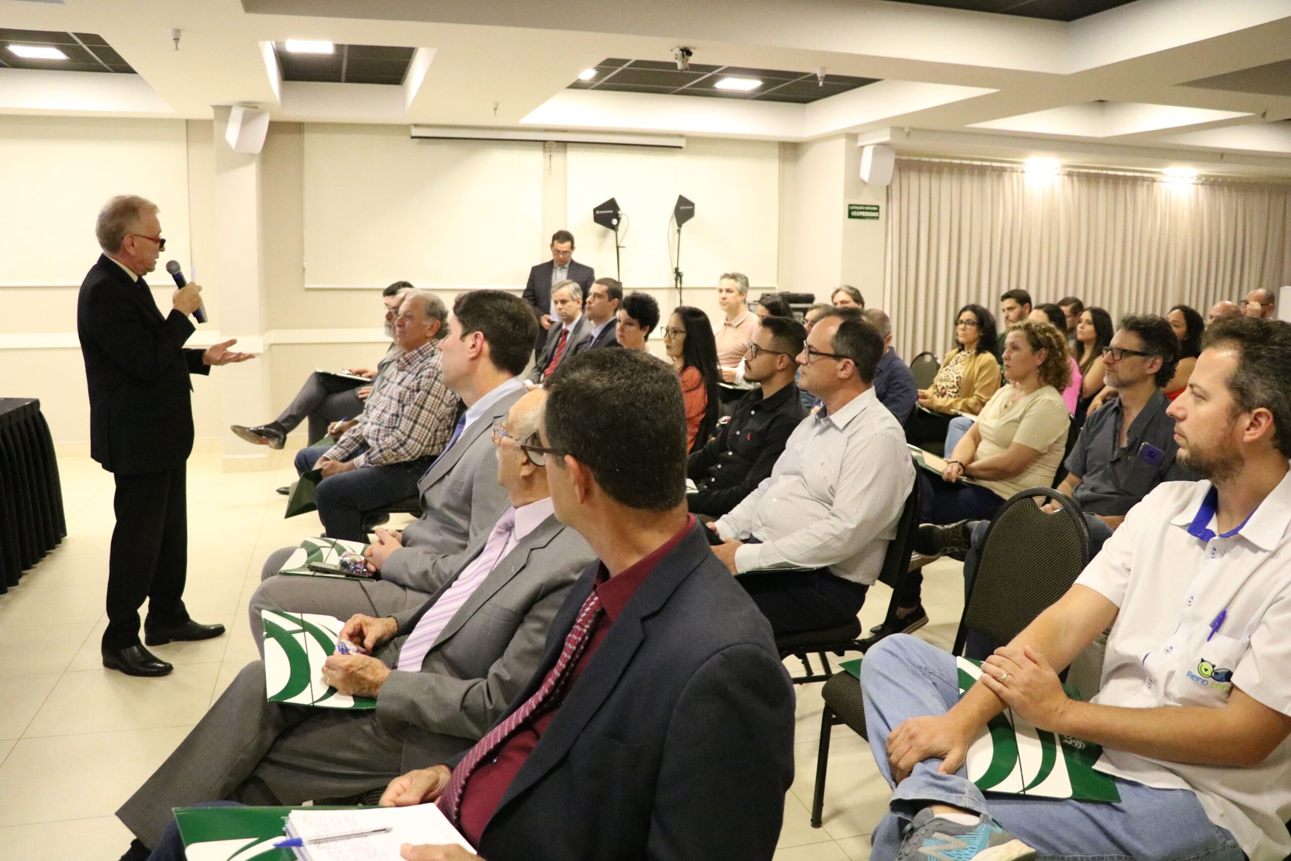 A foto mostra o auditório do CRMV-SP Escuta de Piracicaba lotado de pessoas sentadas em cadeiras com pastas do CRMV-SP verde e brancas no colo. A frente está de pé ao microfone o presidente do CRMV-SP.