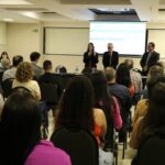 A foto foi tirada da parte de trás do auditório e mostra os participantes sentados em cadeiras de costas para a câmera. A frente estão posicionados atrás de uma mesa com toalha preta e um arranjo de flores, a tesoureira, o presidente e o secretário-geral do CRMV-SP. Eles estão de pé e ao microfone está a tesoureira.