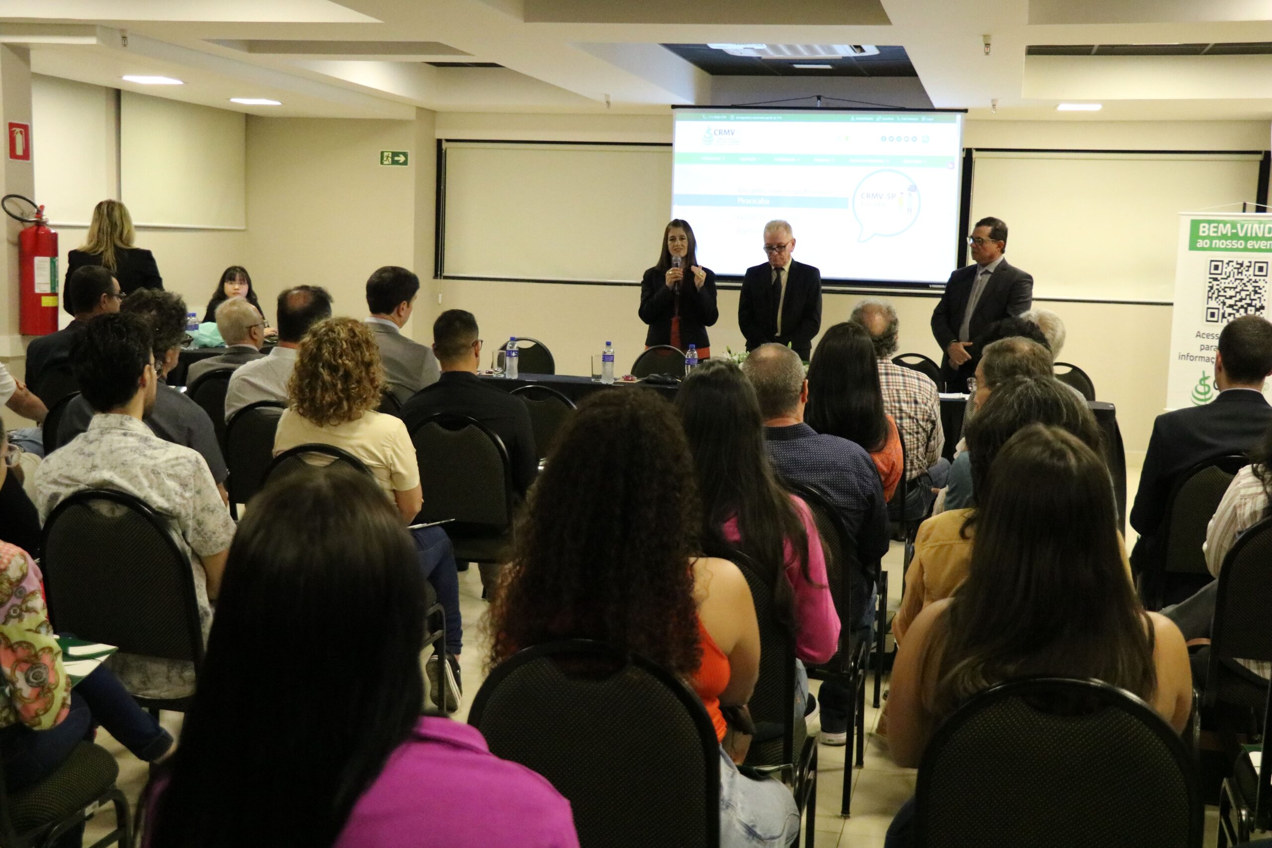 A foto foi tirada da parte de trás do auditório e mostra os participantes sentados em cadeiras de costas para a câmera. A frente estão posicionados atrás de uma mesa com toalha preta e um arranjo de flores, a tesoureira, o presidente e o secretário-geral do CRMV-SP. Eles estão de pé e ao microfone está a tesoureira.