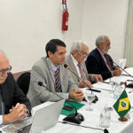 Foto mostra quatro homens sentados. Há uma mesa na frente, com microfones.