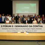 A foto mostra os participantes do Seminário. Eles estão no palco, alguns estão em pé e outros estão sentados. Em frente deles, há um cartaz escrito "II Fórum e I Seminário da Contha".