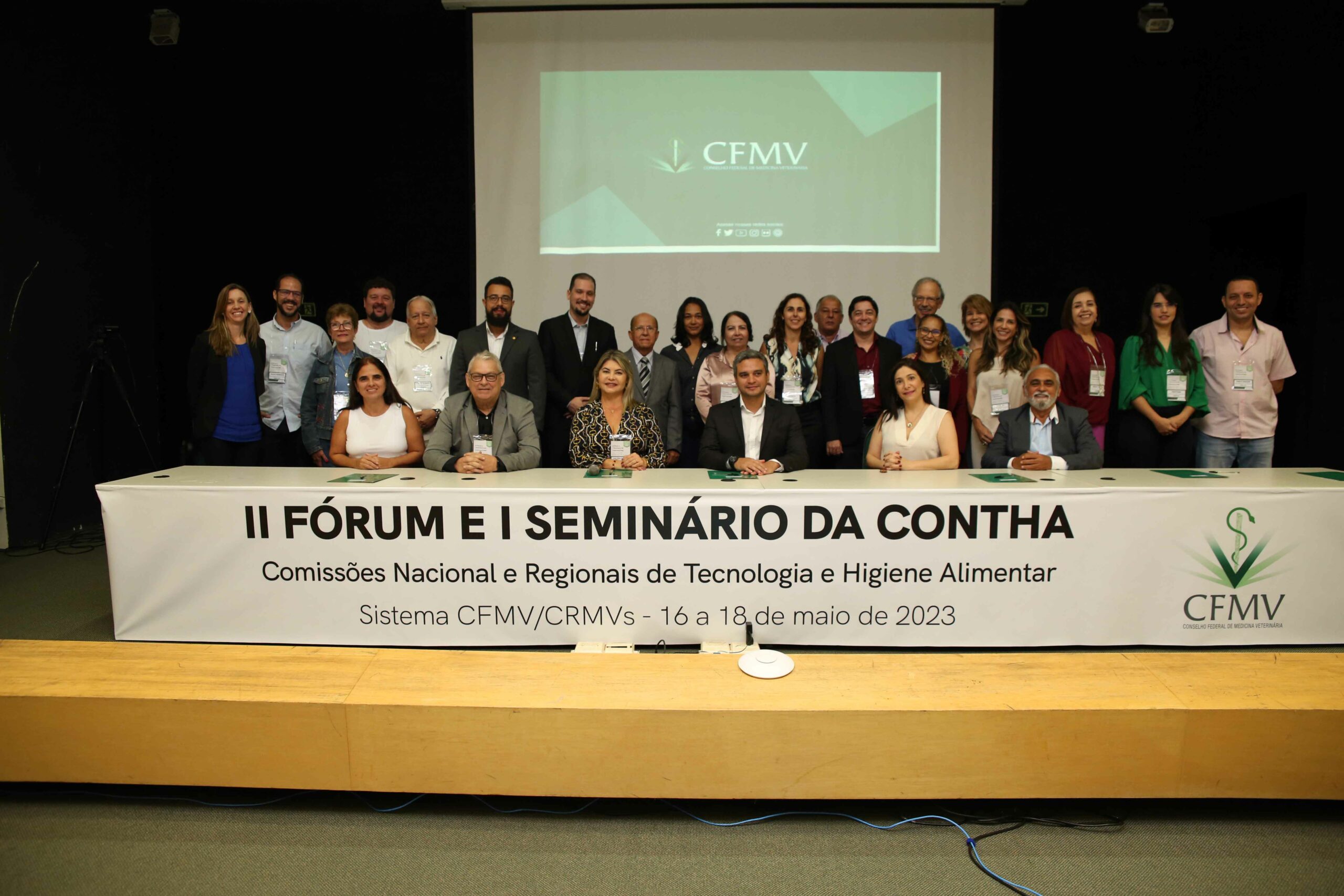 A foto mostra os participantes do Seminário. Eles estão no palco, alguns estão em pé e outros estão sentados. Em frente deles, há um cartaz escrito "II Fórum e I Seminário da Contha".