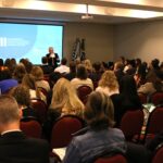 A foto mostra um auditório lotado visto da parte de trás. A frente está de pé ao microfone o presidente do CRMV-SP. Atrás dele um telão com os dizeres: II Encontro de Coordenadores de Cursos do Estado de São Paulo.