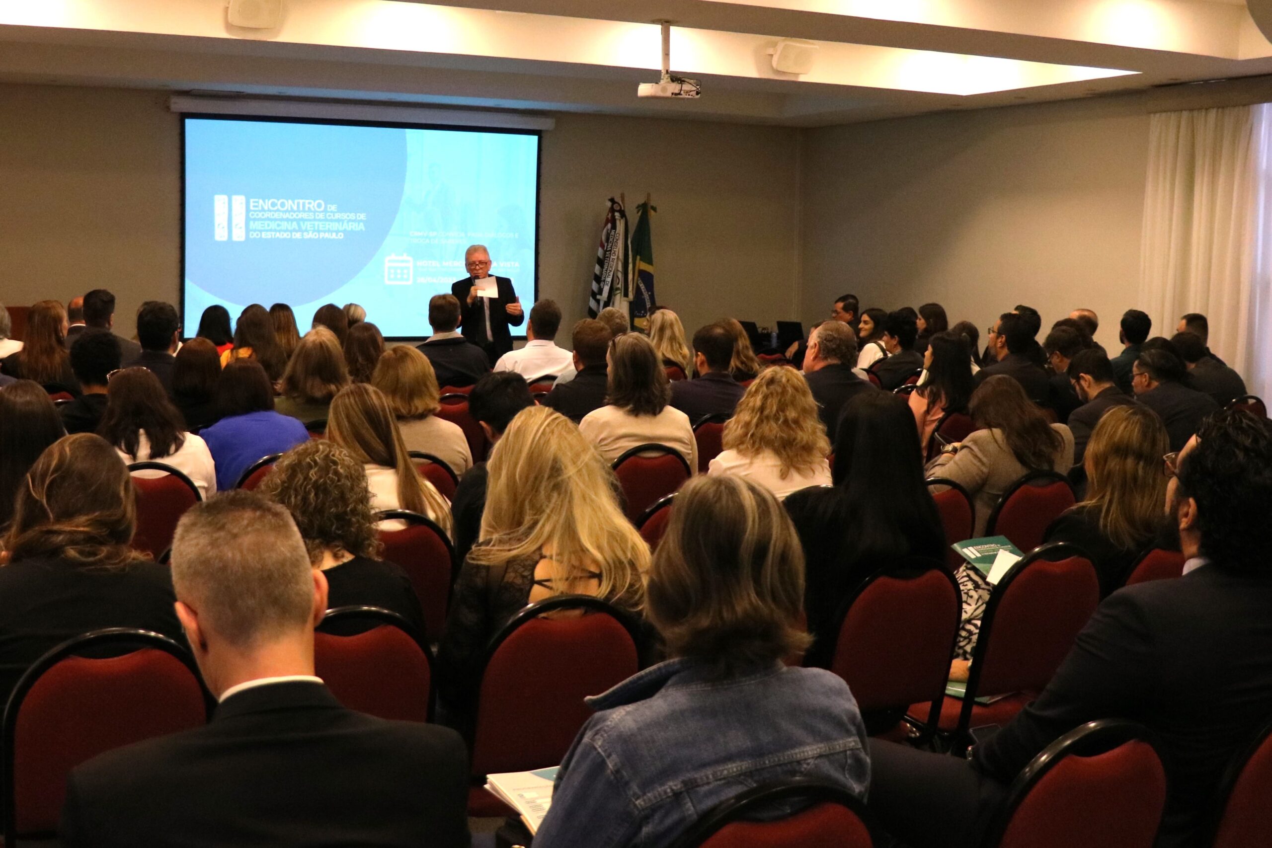 A foto mostra um auditório lotado visto da parte de trás. A frente está de pé ao microfone o presidente do CRMV-SP. Atrás dele um telão com os dizeres: II Encontro de Coordenadores de Cursos do Estado de São Paulo.