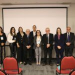 Na foto aparecem representantes dos diversos conselhos regionais do País que compareceram ao evento. Eles estão todos de pé, posando e sorrindo para a câmera, posicionados lado a lado. A frente deles, cadeiras vermelhas do auditório. Atrás deles um telão. Ao todo são seis mulheres e cinco homens.