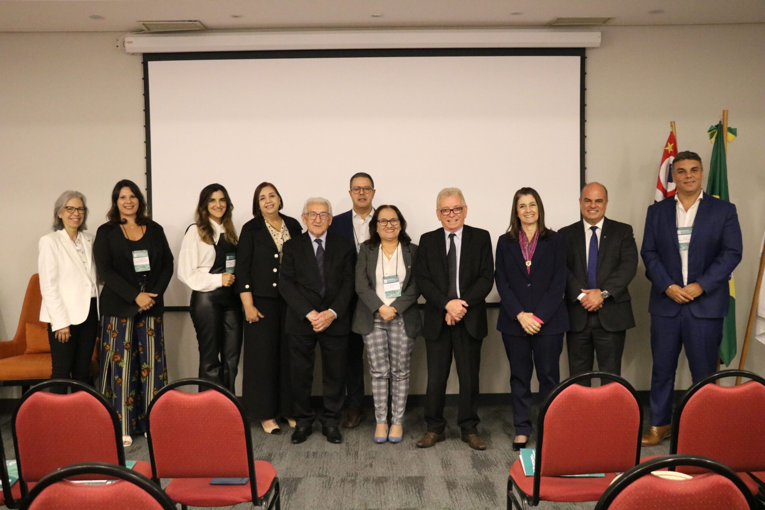 Na foto aparecem representantes dos diversos conselhos regionais do País que compareceram ao evento. Eles estão todos de pé, posando e sorrindo para a câmera, posicionados lado a lado. A frente deles, cadeiras vermelhas do auditório. Atrás deles um telão. Ao todo são seis mulheres e cinco homens.