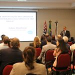 A foto mostra um auditório lotado com pessoas sentadas. Com a posição da câmera, todos estão de cotas voltados para um telão com os dizeres: "Sistema de Certificação de Cursos de Medicina Veterinária do Estado de São Paulo". A frente também o vice-presidente do CRMV-SP, Fabio Manhoso. No ambiente há ainda as bandeiras do estado de São Paulo, do Brasil e do Conselho.