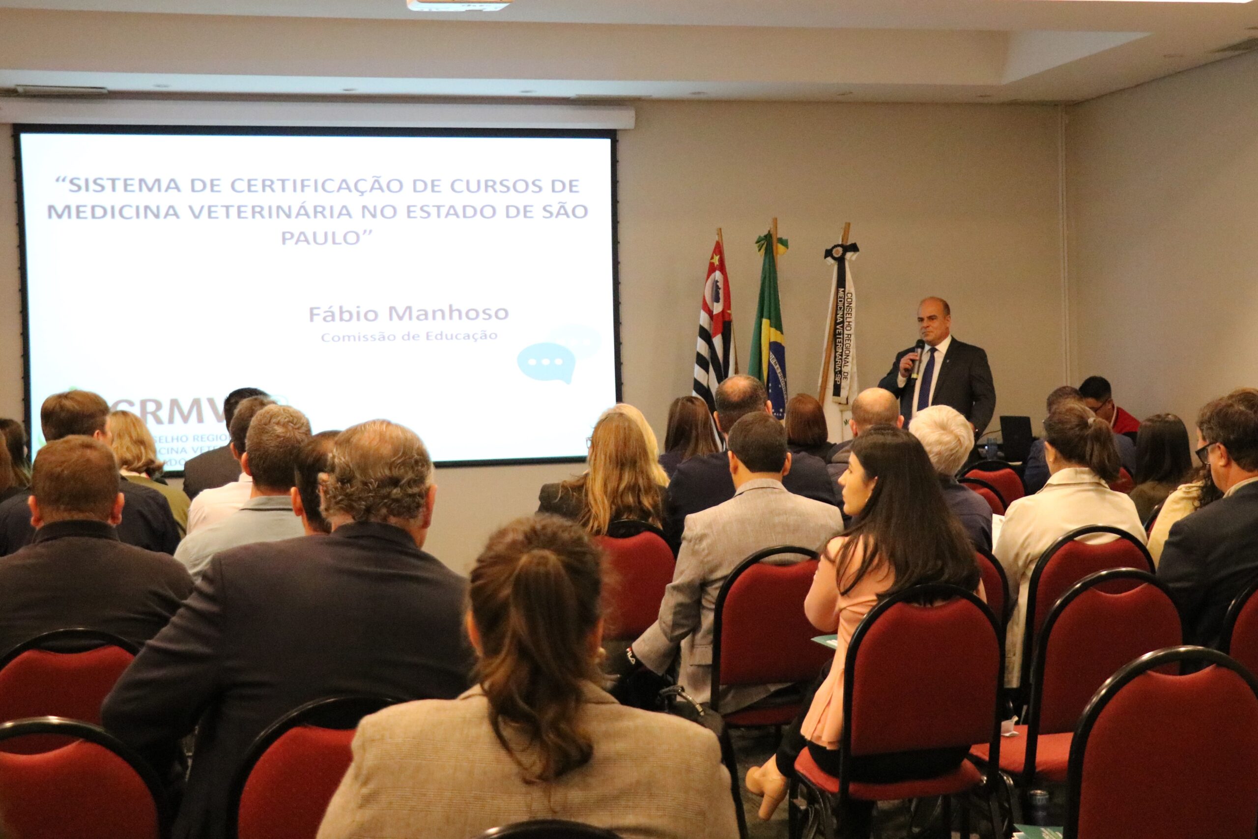 A foto mostra um auditório lotado com pessoas sentadas. Com a posição da câmera, todos estão de cotas voltados para um telão com os dizeres: "Sistema de Certificação de Cursos de Medicina Veterinária do Estado de São Paulo". A frente também o vice-presidente do CRMV-SP, Fabio Manhoso. No ambiente há ainda as bandeiras do estado de São Paulo, do Brasil e do Conselho.