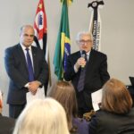 Na foto aparecem em destaque o vice-presidente do CRMV-SP e o presidente do CFMV, o qual fala ao microfone. Eles se dirigem a um auditório lotado. Atrás deles estão as bandeiras do estado de São Paulo, do Brasil e do CRMV-SP.