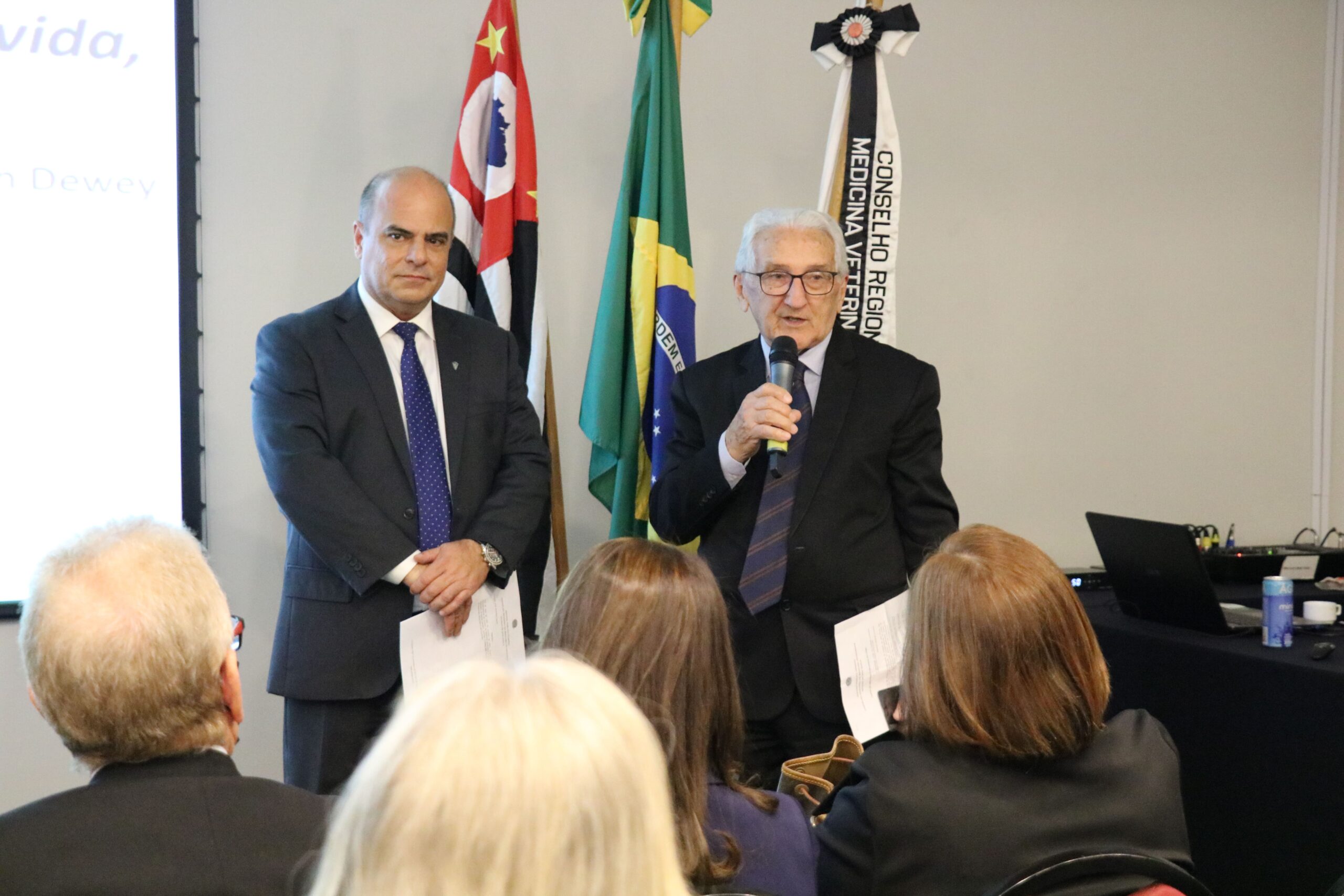 Na foto aparecem em destaque o vice-presidente do CRMV-SP e o presidente do CFMV, o qual fala ao microfone. Eles se dirigem a um auditório lotado. Atrás deles estão as bandeiras do estado de São Paulo, do Brasil e do CRMV-SP.