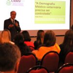 Na foto aparece em foco o consultor ad hc do CRMV-PR, Antônio Felipe Paulino de Figueiredo Wouk. Ele está a frente de um auditório com pessoas na plateia. Ele é um homem franzino de óculo e veste terno. Atrás dele está um telão com a logomarca do CRMV-SP e os dizeres: Demografia da Medicina Veterinária no Brasil".