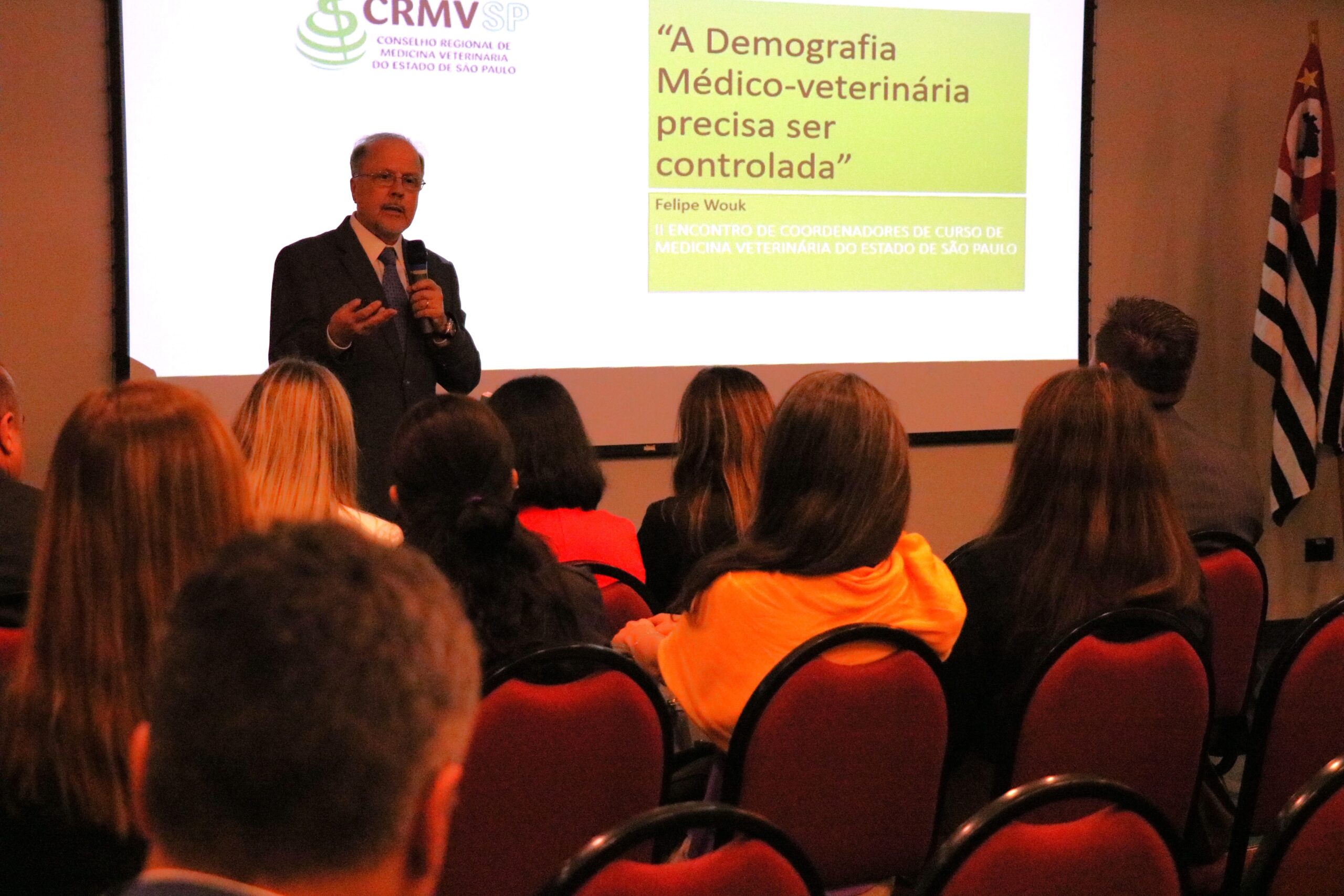 Na foto aparece em foco o consultor ad hc do CRMV-PR, Antônio Felipe Paulino de Figueiredo Wouk. Ele está a frente de um auditório com pessoas na plateia. Ele é um homem franzino de óculo e veste terno. Atrás dele está um telão com a logomarca do CRMV-SP e os dizeres: Demografia da Medicina Veterinária no Brasil".