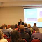 Na foto aparece metade da sala de um auditório com pessoas sentadas em cadeiras vermelhas. Eles estão de costas para a câmera. A frente está o palestrante Adolfo-Ignácio Calderón ao microfone. Atrás dele há um telão com a projeção de caixas coloridas com os dizeres: "Avaliação Externa e Rankings acadêmicos na educação superior.