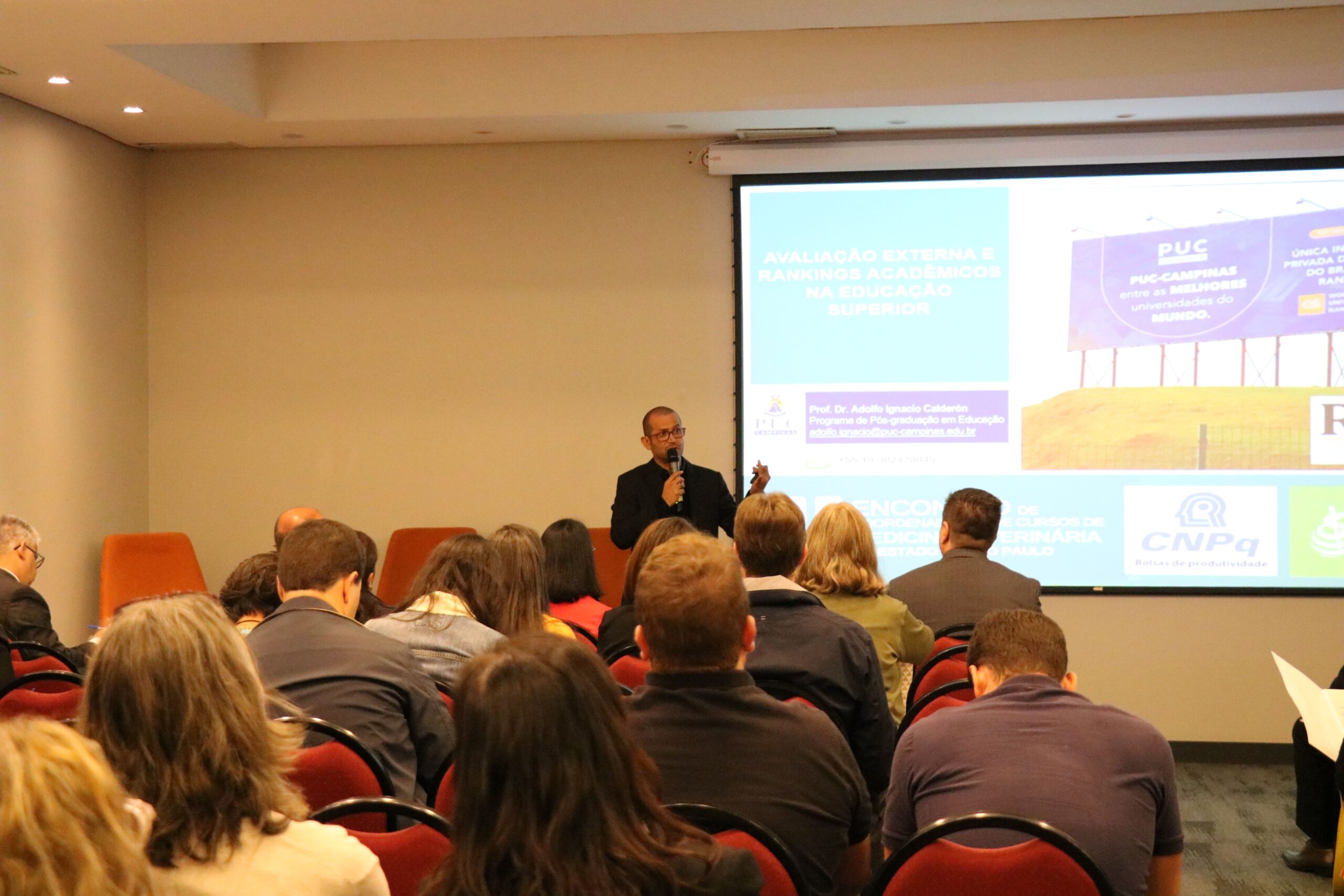 Na foto aparece metade da sala de um auditório com pessoas sentadas em cadeiras vermelhas. Eles estão de costas para a câmera. A frente está o palestrante Adolfo-Ignácio Calderón ao microfone. Atrás dele há um telão com a projeção de caixas coloridas com os dizeres: "Avaliação Externa e Rankings acadêmicos na educação superior.