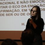 Na imagem está a assessora pedagógica da Unesp Botucatu, Eliana Cordeiro Curvelo. Ela está na sombra emitida pelo projetor e na imagem destaca-se apenas seu rosto. Ela é uma mulher branca de cabelos longos e usa óculos. Atrás dela há um telão com a projeção de um slide branco com frases na cor preta. A foto mostra apenas parte da tela.