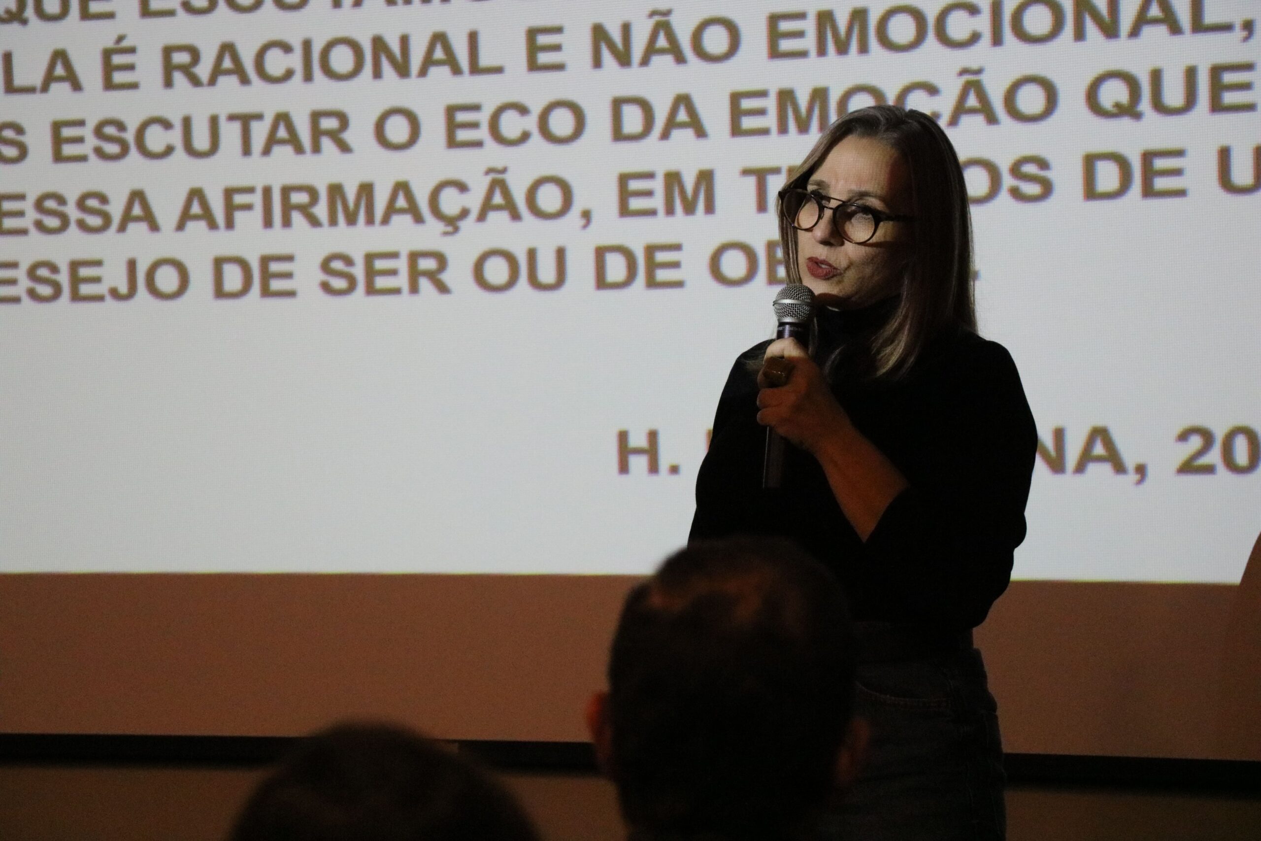 Na imagem está a assessora pedagógica da Unesp Botucatu, Eliana Cordeiro Curvelo. Ela está na sombra emitida pelo projetor e na imagem destaca-se apenas seu rosto. Ela é uma mulher branca de cabelos longos e usa óculos. Atrás dela há um telão com a projeção de um slide branco com frases na cor preta. A foto mostra apenas parte da tela.