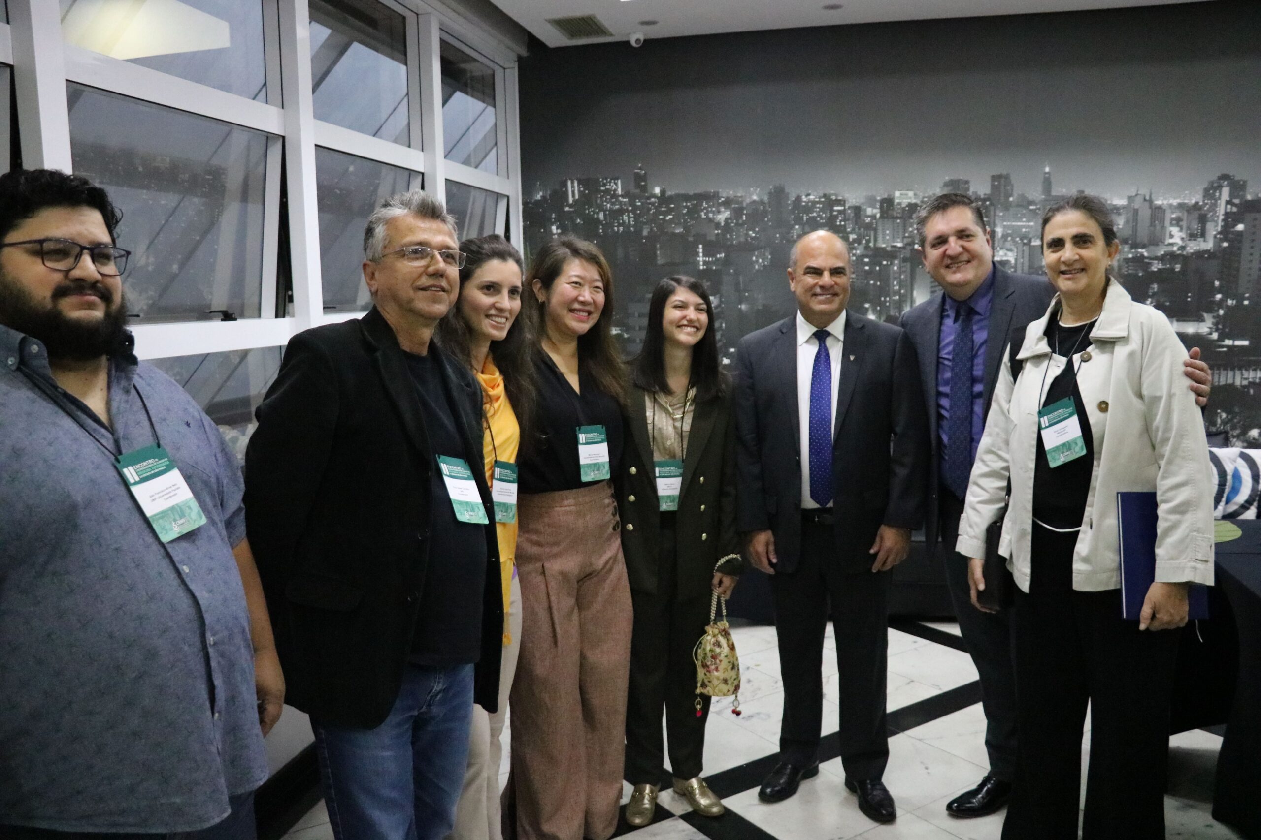 Na foto há quatro homens e quatro mulheres, coordenadores de instituições de ensino superior, representantes da ABHV e do CRMV-SP. Todos estão de pé, sorriem para a câmera e estão posicionados lado a lado a frente de um painel em preto e branco da cidade de São Paulo.