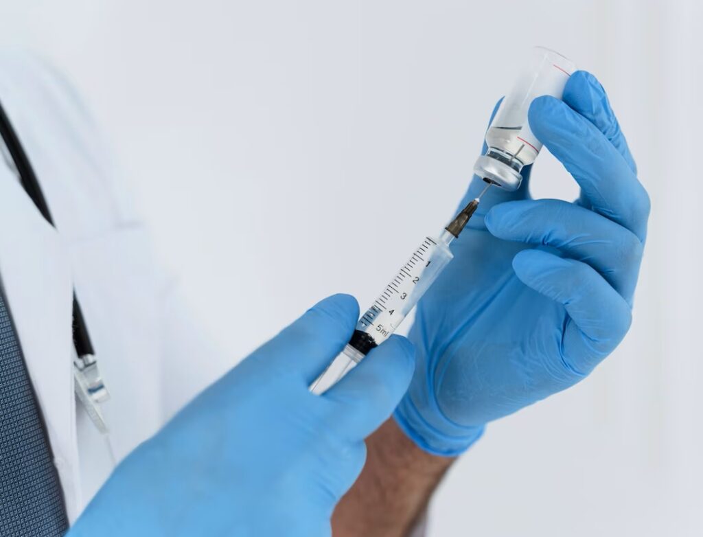 Imagem mostra profissional da saúde segurando uma seringa que está extraindo líquido de um pequeno frasco.