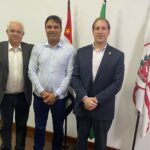 Na imagem aparecem três homens de pé, lado a lado, olhando e sorrindo para a câmera. Ao fundo três bandeiras: do estado de São Paulo, do Brasil e do município de São Paulo. Eles são o assessor de Gabinete do CRMV-SP, o secretário adjunto da Casa Civil, e o presidente da Comissão Técnica de Políticas Públicas, respectivamente.