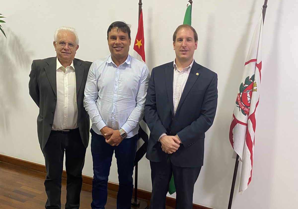 Na imagem aparecem três homens de pé, lado a lado, olhando e sorrindo para a câmera. Ao fundo três bandeiras: do estado de São Paulo, do Brasil e do município de São Paulo. Eles são o assessor de Gabinete do CRMV-SP, o secretário adjunto da Casa Civil, e o presidente da Comissão Técnica de Políticas Públicas, respectivamente.