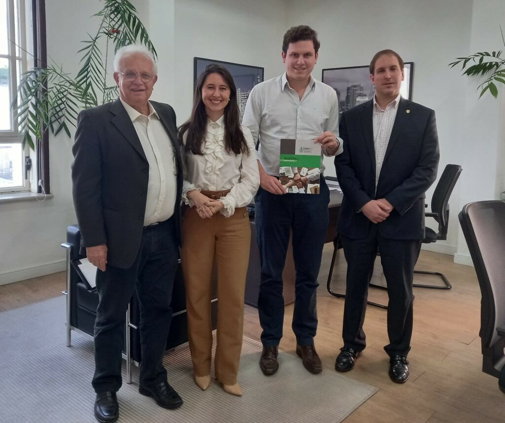 Na imagem estão quatro pessoas, sendo uma mulheres e três homens. Estão todos de pé, olhando e sorrindo para a foto. São eles: o assessor de Gabinete do CRMV-SP, a coordenadora de relacionamento com entidades, o secretário de Relações Institucionais da Prefeitura de São Paulo, e o presidente da Comissão Técnica de Políticas Públicas. O secretário está mais ao centro e segura um relatório de gestão do CRMV-SP.