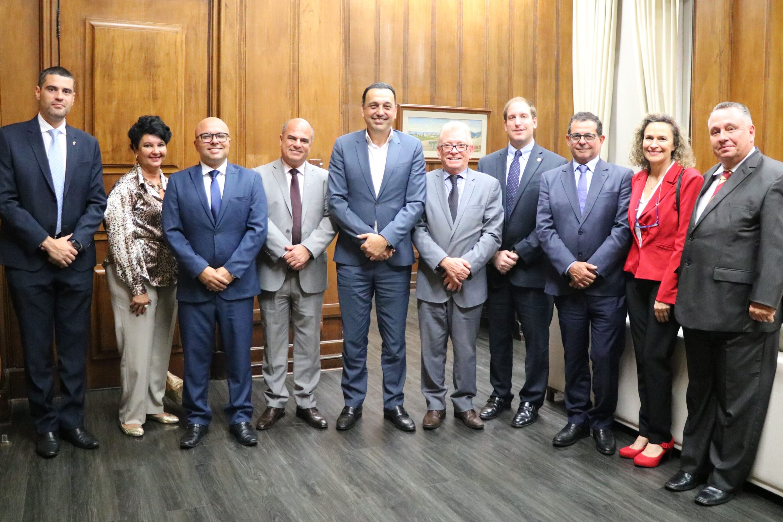 Na imagem estão nove representantes do CRMV-SP, sendo duas mulheres e sete homens, e o vice-governador de São Paulo. Estão todos de pé, lado a lado, e sorriem para a câmera. Ao fundo uma parede de madeira. Eles estão na sala do Gabinete do vice-governador.