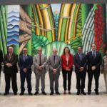 Na foto os nove representantes do CRMV-SP estão de pé, dispostos lado a lado, em frente a um painel artístico que ocupa toda a parede do hall do Palácio dos Bandeirantes. O tema é natureza e ele é bastante colorido, trazendo as cores azul, verde, amarelo e laranja em destaque. Todos sorriem para a câmera.