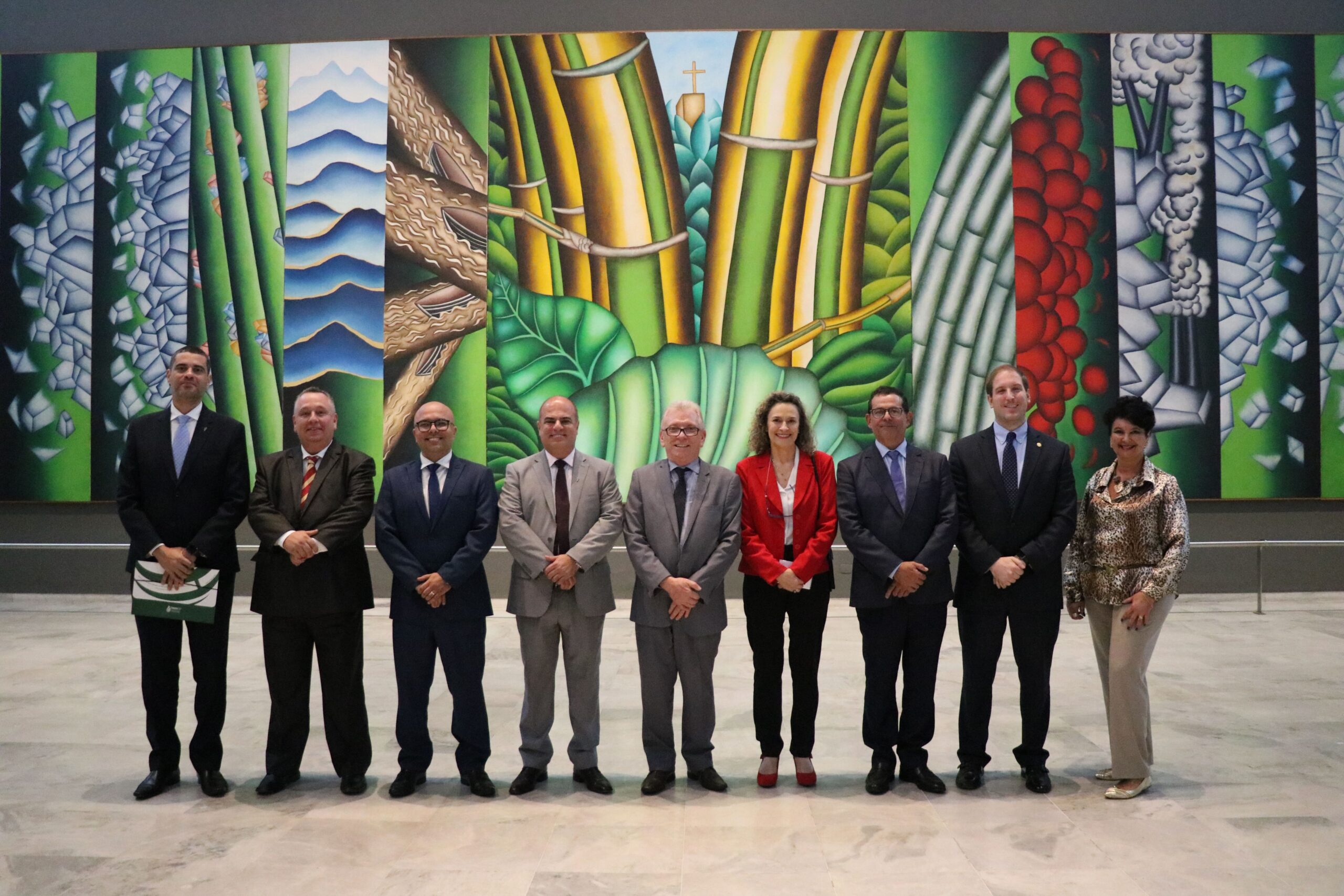 Na foto os nove representantes do CRMV-SP estão de pé, dispostos lado a lado, em frente a um painel artístico que ocupa toda a parede do hall do Palácio dos Bandeirantes. O tema é natureza e ele é bastante colorido, trazendo as cores azul, verde, amarelo e laranja em destaque. Todos sorriem para a câmera.