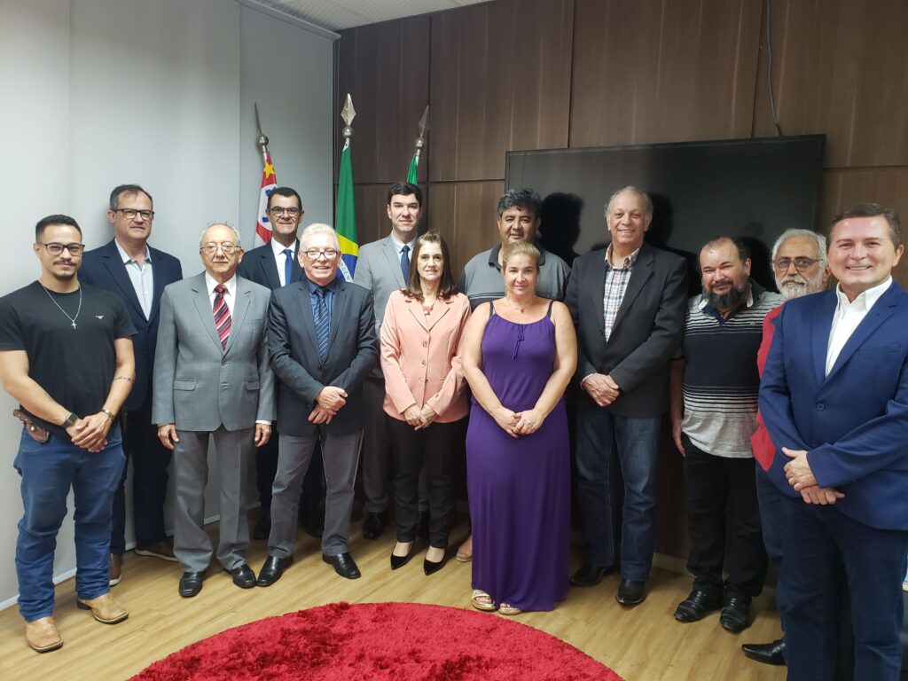 Na foto estão todos os representantes do CRMV-SP e da associação de médicos-veterinários de Piracicaba, o diretor do DBEA, o presidente da Câmara Municipal de Piracicaba e vereadores. Estão todos de pé posando e sorrindo para a câmera. São 11 homens e duas mulheres no total. Na sala há um tapete vermelho redondo a frente deles, e bandeiras do Brasil, do estado de São Paulo e do município atrás deles, assim como uma televisão desligada.