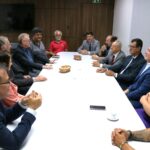 Na foto aparecem todos os representantes do Conselho, da associação de médicos-veterinários de Piracicaba, do DBEA e da Câmara Municipal de Piracicaba. São 11 homens e duas mulheres no total. les estão sentados ao entorno de uma mesa de cor branca atentos a fala do presidente da casa legislativa.
