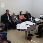 Representantes do CRMV-SP e da associação de médicos-veterinários de Piracicaba estão sentados em sofás e cadeiras na sala do delegado seccional de Piracicaba, o qual está atrás de mesa cheia de papéis. Todos estão atentos a fala do delegado.