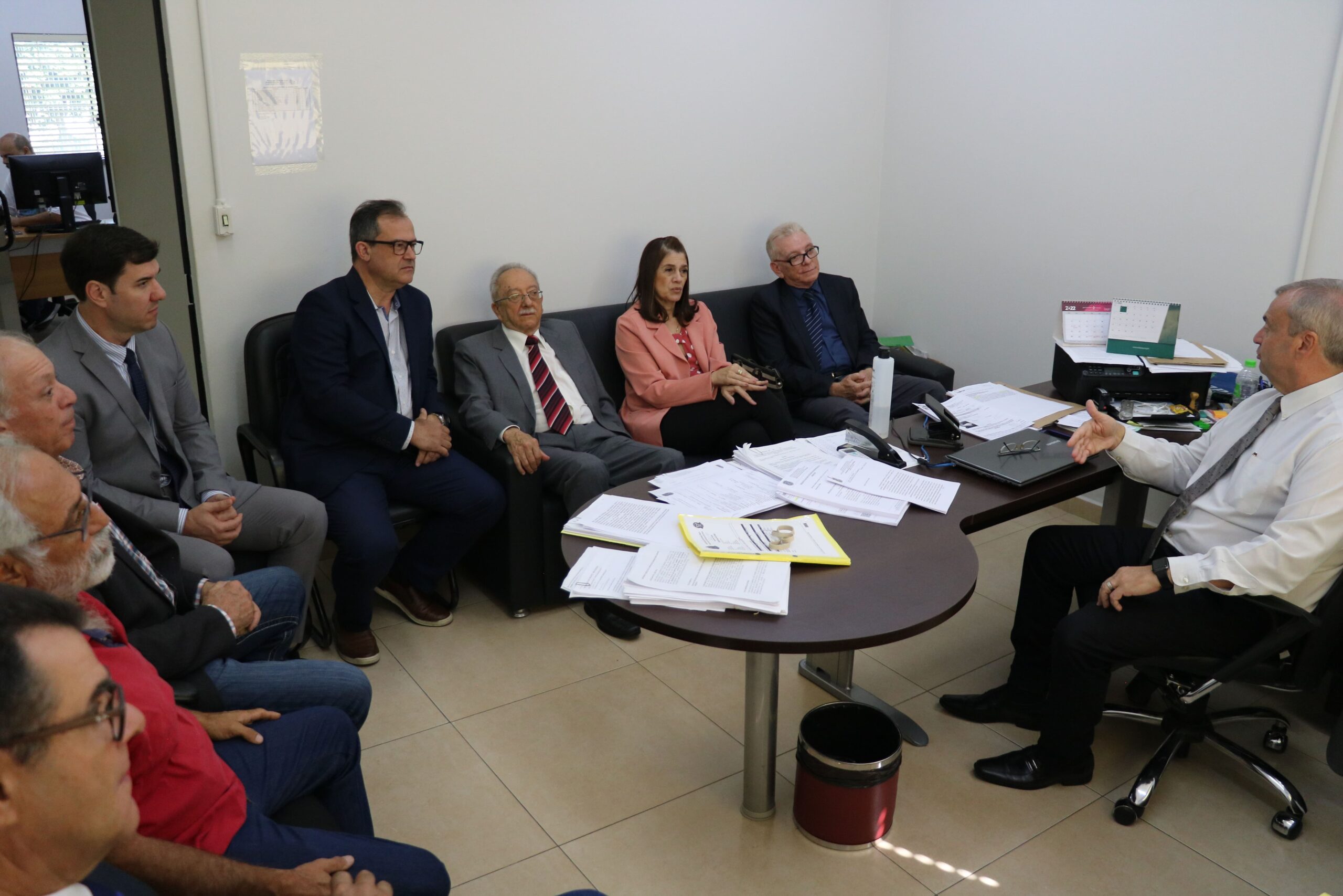 Representantes do CRMV-SP e da associação de médicos-veterinários de Piracicaba estão sentados em sofás e cadeiras na sala do delegado seccional de Piracicaba, o qual está atrás de mesa cheia de papéis. Todos estão atentos a fala do delegado.